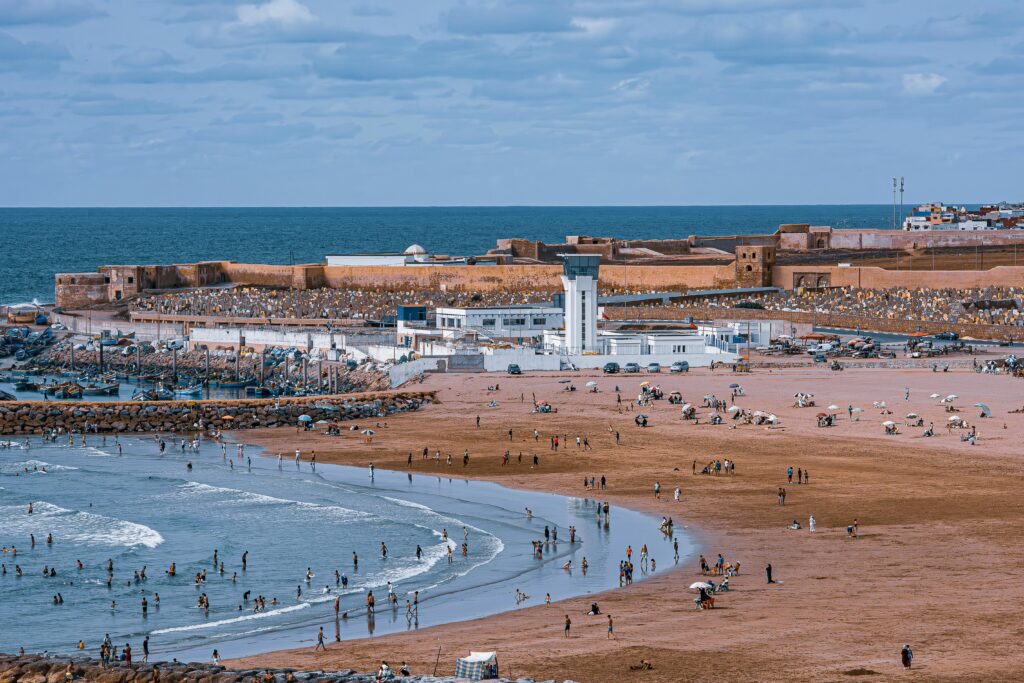 A vibrant beach scene near Rabat's historic fort with people enjoying sun and sea.